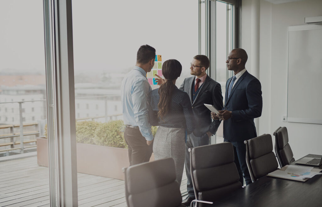 A group of people in business dress standing next to a tall meeting room window with bright sunlight outside